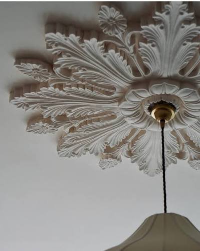 Balta lubų rozetė su lapų ir gėlių ornamentais aplink pakabinamo šviestuvo tvirtinimo vietą